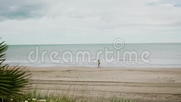 穿着黄色夹克和牛仔裤短裤的年轻女孩沿着地中海海滩散步视频的预览图
