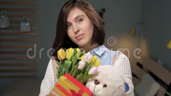 女孩带着礼物迷人的年轻女子独自在房间里手里拿着一束鲜花礼物软玩具看着视频的预览图