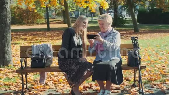 美丽的金发女孩年长的女人坐在长凳上看着智能手机上的东西视频的预览图
