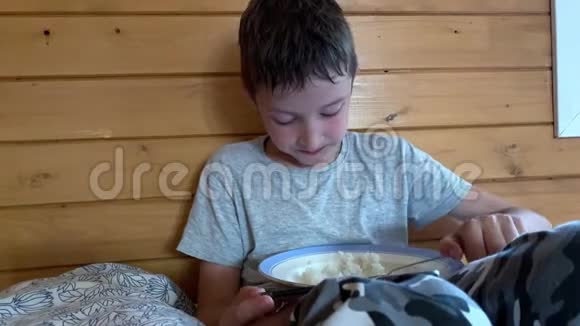 一个非常饥饿的男孩头发湿漉漉的坐在农村木屋的床上贪婪地吃着米饭他在外面玩耍视频的预览图