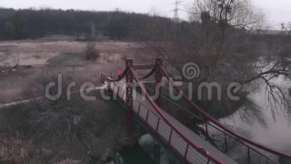 空中无人机观看女子运动员慢跑过桥穿橙色夹克的女人在公园里和河和桥一起跑体育项目视频的预览图