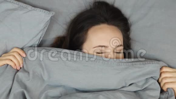 年轻女子躺在床上看着相机肖像俯视迷人的黑发女孩躺在铺着灰色毯子的床上视频的预览图