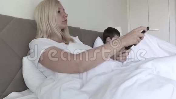 一个金发女人躺在床上通过遥控器切换频道她的儿子在后台玩智能手机游戏视频的预览图