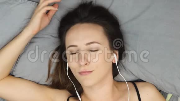 成年女孩躺在床上听着耳机里的音乐看着相机俯视图戴着耳机的布鲁内特女士听视频的预览图