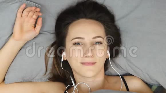 幸福的年轻女子在耳机听音乐在床上俯视白种人黑发女性躺在床上的肖像听视频的预览图