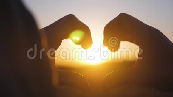 年轻美丽的女孩用她的手在心形框架夕阳日落时使心快乐的情感观念视频的预览图