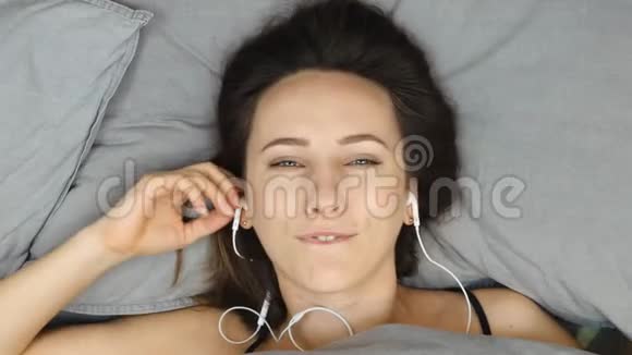 穿着黑色内衣的迷人害羞女人躺在灰色的床上听着耳机里的音乐年轻的黑发女孩在耳机里撒谎视频的预览图