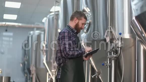 有胡子的男性啤酒商站在啤酒罐附近用平板电脑捕捉读数手工艺啤酒视频的预览图