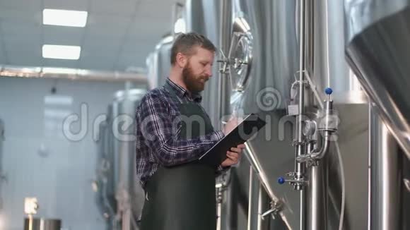 有胡子的男酿酒师布鲁塔站在啤酒罐附近记录仪器读数手工艺啤酒生产视频的预览图