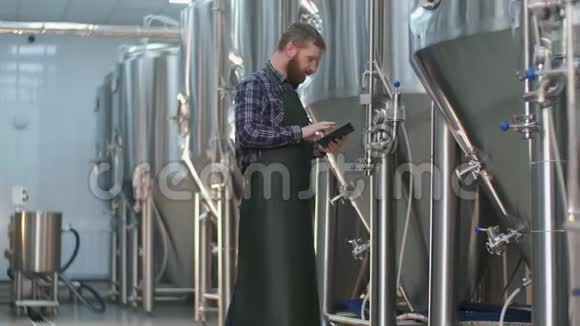 一个有胡子的男性啤酒师用平板电脑监控啤酒罐的读数手工艺啤酒生产视频的预览图
