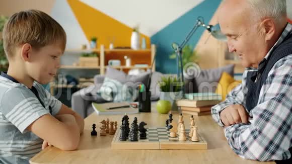聪明的孩子在家里和爷爷下棋享受室内游戏视频的预览图