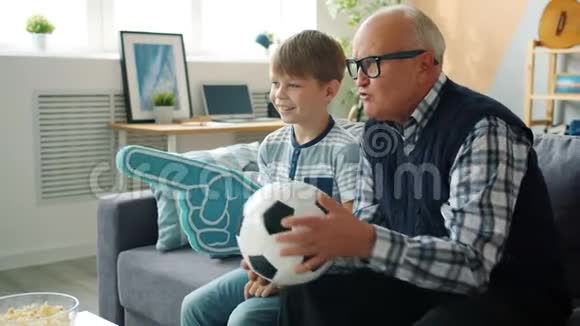 可爱的爷爷和快乐的男孩在电视上捧着泡沫的手和球看足球视频的预览图