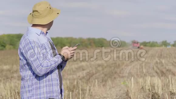 英俊的农民与智能手机站在田间向日葵与联合收割机的背景现代概念视频的预览图