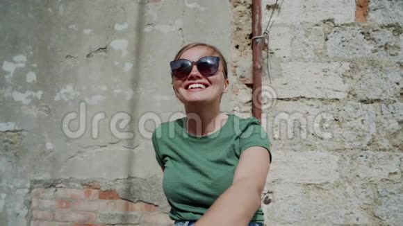 戴眼镜的漂亮女孩的户外肖像坐在老砖墙附近微笑视频的预览图