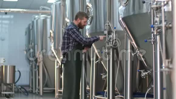 一个长着胡须的男性啤酒师用平板电脑检查啤酒罐上设备的读数手工艺啤酒生产视频的预览图