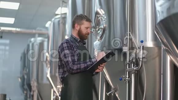 一个穿着带胡须的围裙的男性啤酒商站在啤酒罐附近写下读数手工艺啤酒生产视频的预览图