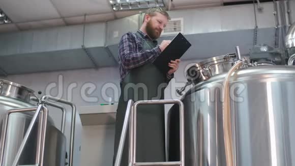 一个穿着带胡须的围裙的男性啤酒商站在啤酒酿造罐附近正在阅读生产视频的预览图