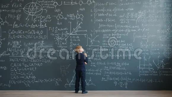 穿西装的体贴孩子在学校上课时拿着公式走在黑板前思考视频的预览图