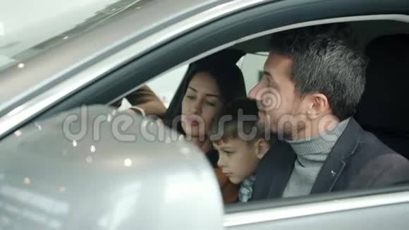 幸福的家庭和可爱的孩子坐在车里在展厅里和推销员交谈视频的预览图