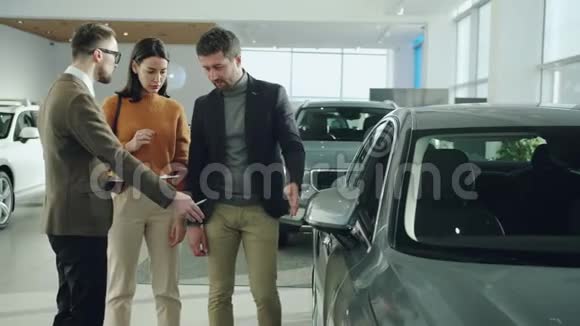 自信的推销员在展示汽车的汽车陈列室里和快乐的年轻夫妇交谈视频的预览图