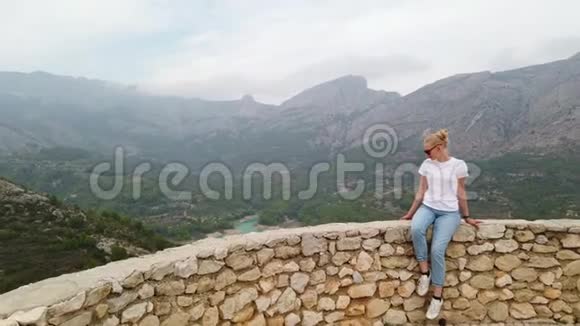 女人坐着看着一个美丽的风景从一座古老城堡的墙上看到一个蓝色的湖和山视频的预览图