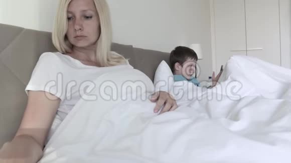 一个金发女人躺在床上通过遥控器切换频道她的儿子在后台玩智能手机游戏视频的预览图