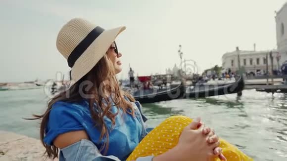 可爱的女孩戴着太阳镜帽子坐在码头上和游客一起看着贡多拉斯视频的预览图
