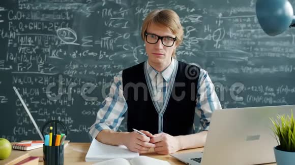 戴眼镜的严肃的年轻人看着摄像机坐在教室的桌子上的画像视频的预览图
