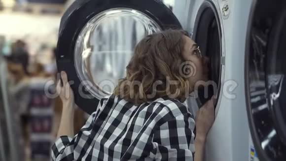 一个年轻的积极的女人穿着格子衬衫在家用电器商店里选择洗衣机往里面看视频的预览图