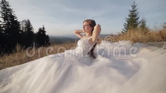 美丽可爱的新娘穿着婚纱坐在山坡上的草地上视频的预览图