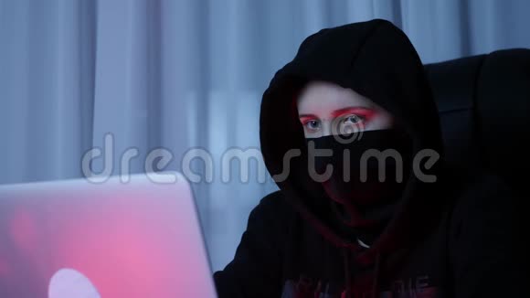 在电脑上关闭戴黑色面罩和引擎盖的女性面部密码黑客的脸看着笔记本电脑屏幕和编码网络视频的预览图