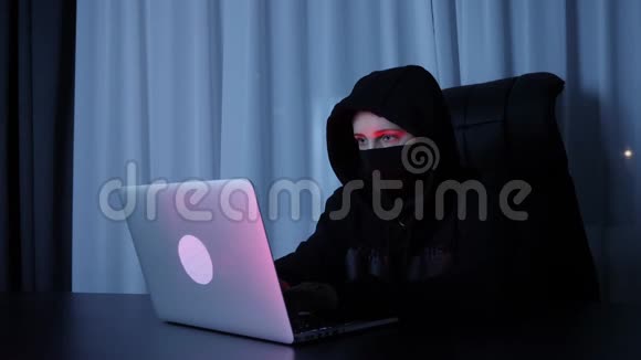 女黑客坐在黑暗的房间里在笔记本电脑键盘上打字戴着黑色面具和罩的女人在屏幕上打电话视频的预览图