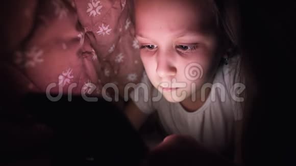 一个年轻的小女孩在睡觉后很晚躲在毯子下使用数字平板智能手机设备孤独视频的预览图