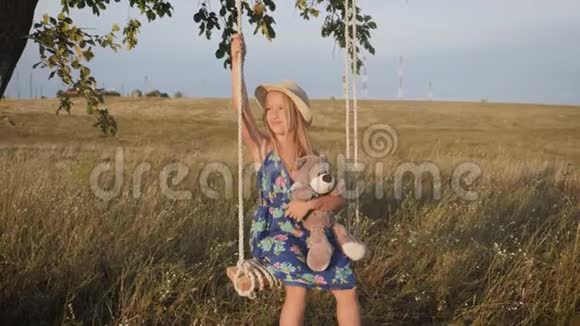 在日落时戴帽子的快乐有趣的女孩正在玩一只泰迪熊在绳子上摆动孩子在户外玩大自然视频的预览图