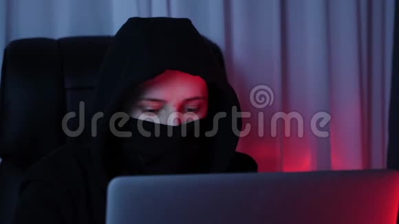女人脸上戴着黑色面具和帽衫看着电脑屏幕打破密码年轻黑客破坏了我的安全保护视频的预览图