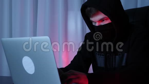 黑客脸戴着黑色面具和引擎盖看着坐在黑暗房间里的笔记本电脑屏幕穿黑色帽衫的男性c编码视频的预览图