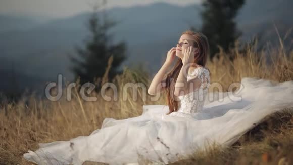 美丽可爱的新娘穿着婚纱坐在山坡上的草地上视频的预览图
