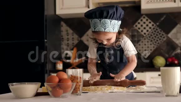 一个穿着厨师帽和围裙可爱的小女孩在家里卷着一个擀面杖面团视频的预览图
