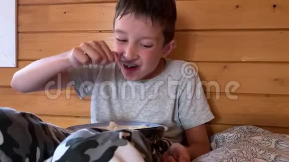 一个非常饥饿的男孩头发湿漉漉地坐在乡村木屋的床上贪婪地吃着米饭他玩着视频的预览图