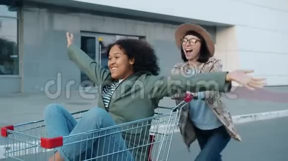 快乐的女孩朋友骑着购物车在商场附近的户外大笑视频的预览图