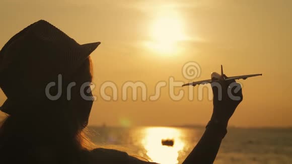 剪影女孩在橙色日落时在海上玩玩具飞机手拿小飞机特写概念旅行视频的预览图