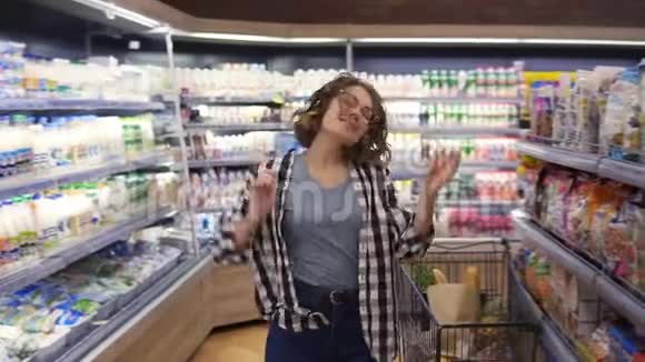 在超市快乐的年轻女孩在超市的货架之间滑稽地跳舞穿牛仔裤和黑色的卷发女孩视频的预览图
