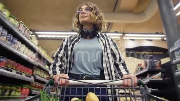 穿着格子衬衫和戴着耳机的开朗女人走在杂货店里用食物操纵购物车视频的预览图