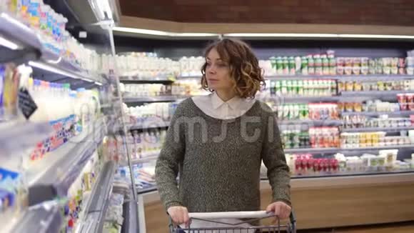 在超市里走着走着的迷人的年轻女人停在靠近冰箱的架子上选择买一个视频的预览图