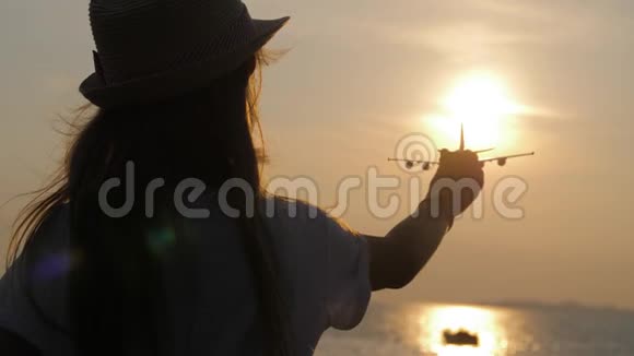 剪影女孩在橙色日落时在海上玩玩具飞机手拿小飞机特写概念旅行视频的预览图