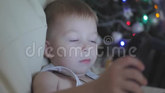 小男孩带着平板电脑在黑暗的房间里在背景圣诞树的夜晚明亮的彩色光线视频的预览图