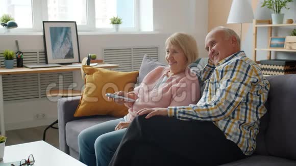 年长的男人和女人在公寓的沙发上看电视和聊天视频的预览图
