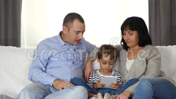 一家人坐在沙发上全神贯注地看着平板电脑屏幕爸爸妈妈还有一个小孩视频的预览图