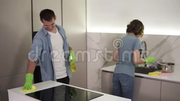 年轻微笑的已婚夫妇穿着休闲服装搬到新家后两人都戴着绿色手套打扫厨房视频的预览图