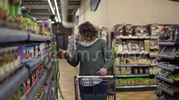 白种人妇女的正面景色是拿着一辆手推车走在商店货架旁在杂货店里挑选一个玻璃瓶然后把它放在视频的预览图